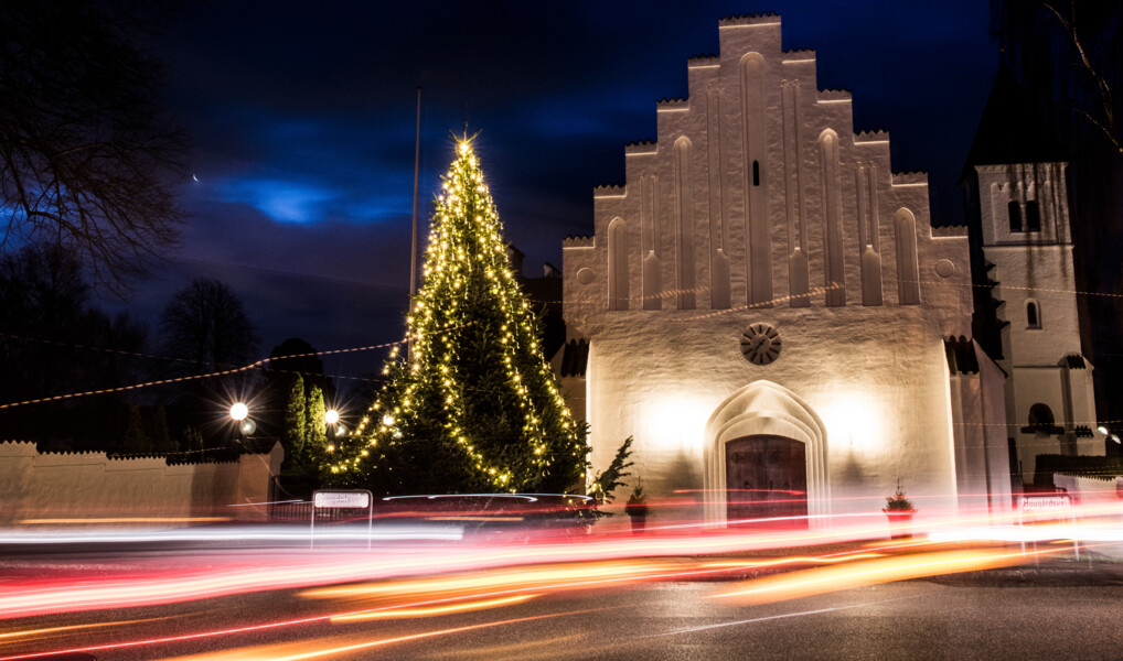 Et juletræ står oplyst foran en landsbykirke